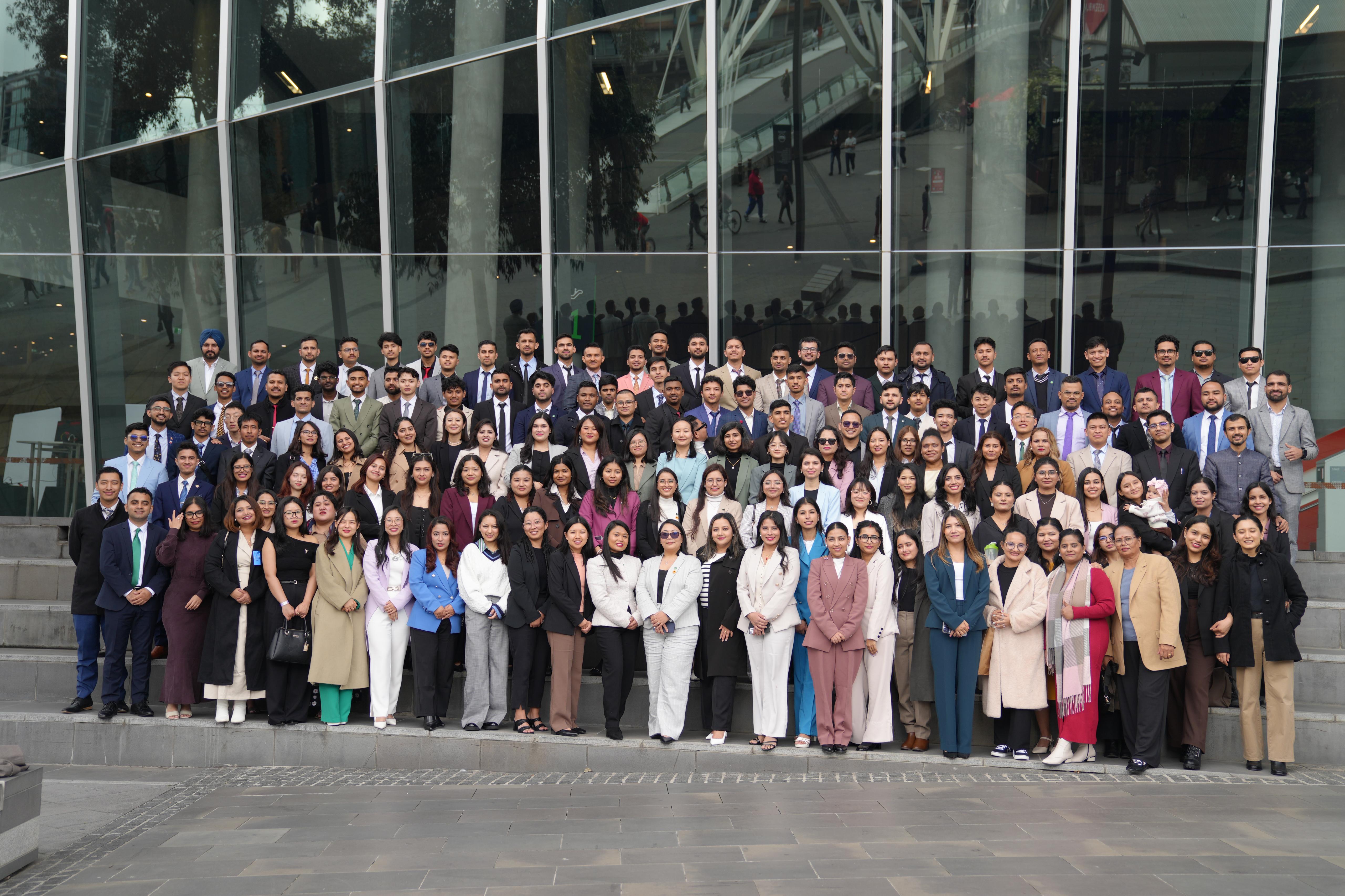 Large professional group photo of Stellars Group members in business attire standing in front of a modern glass building, showcasing the diverse and accomplished community of professionals and students